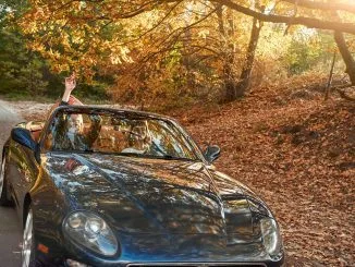 A black roofless car driving fast on the countryside asphalt road against morning sky with a beautiful sunrise. Autumn season, falling leaves
